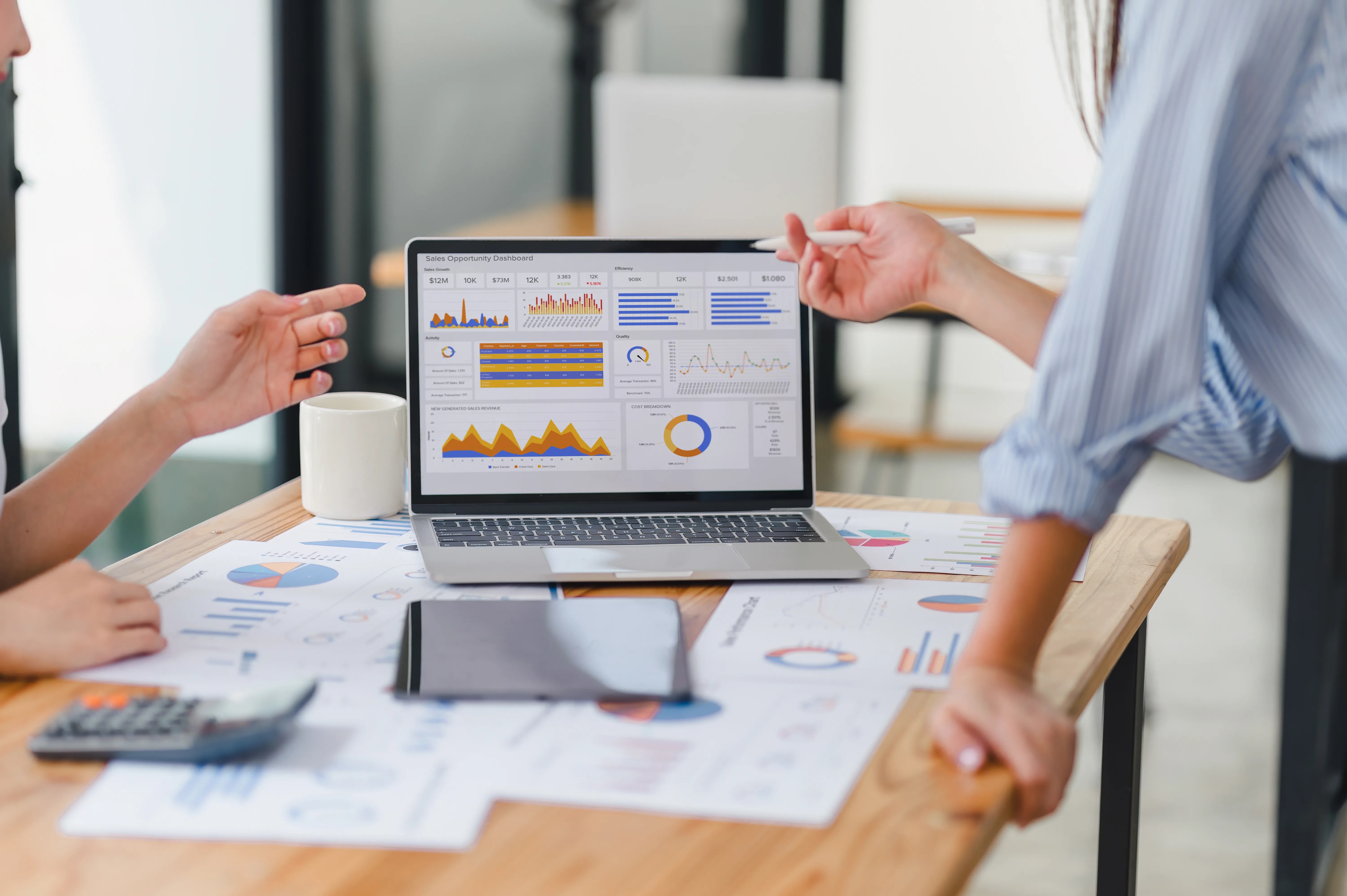Two colleagues discuss a sales dashboard displayed on a laptop surrounded by printed charts
