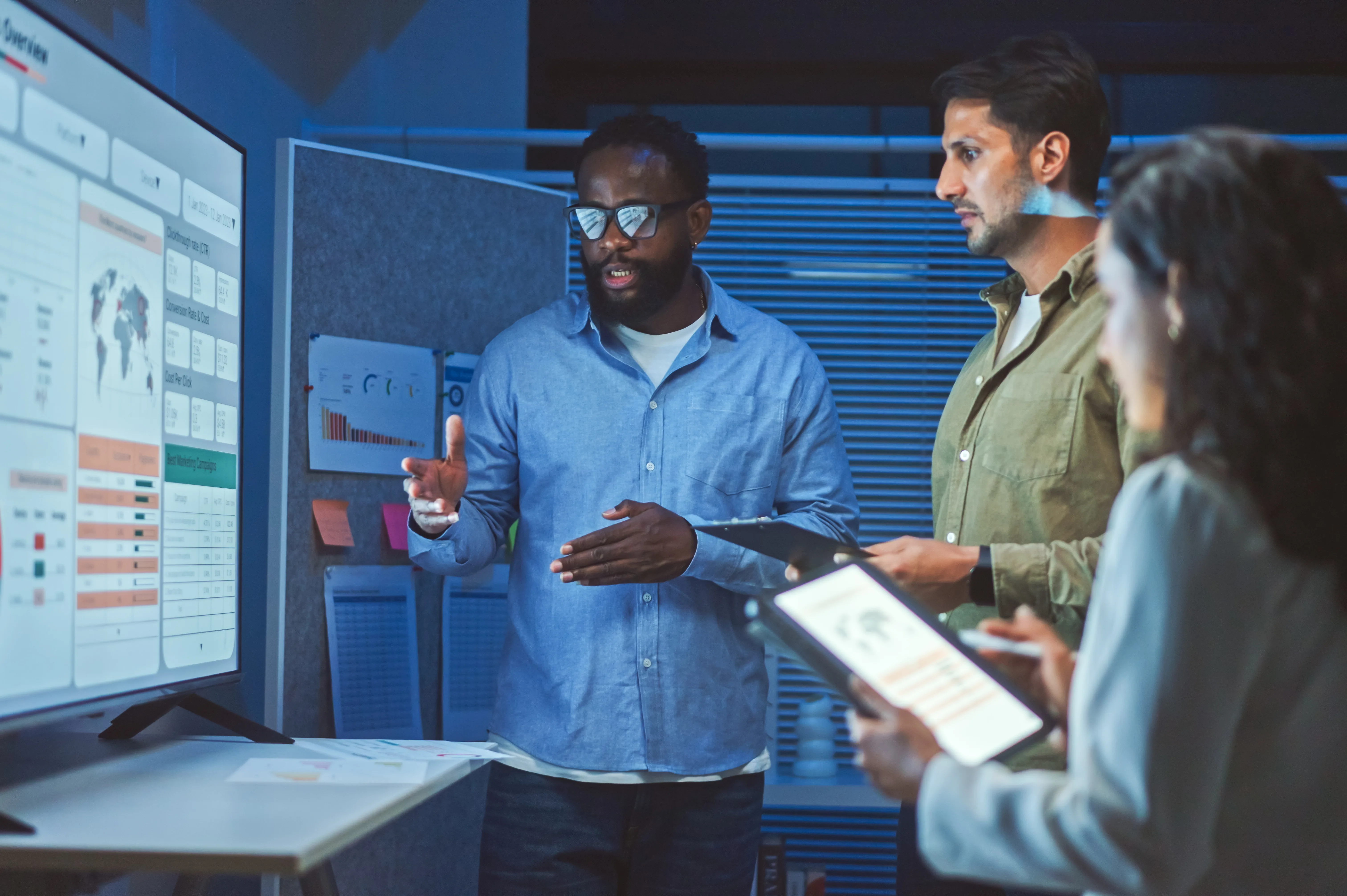 Man presents a data dashboard on a wall screen to two colleagues in a dim office