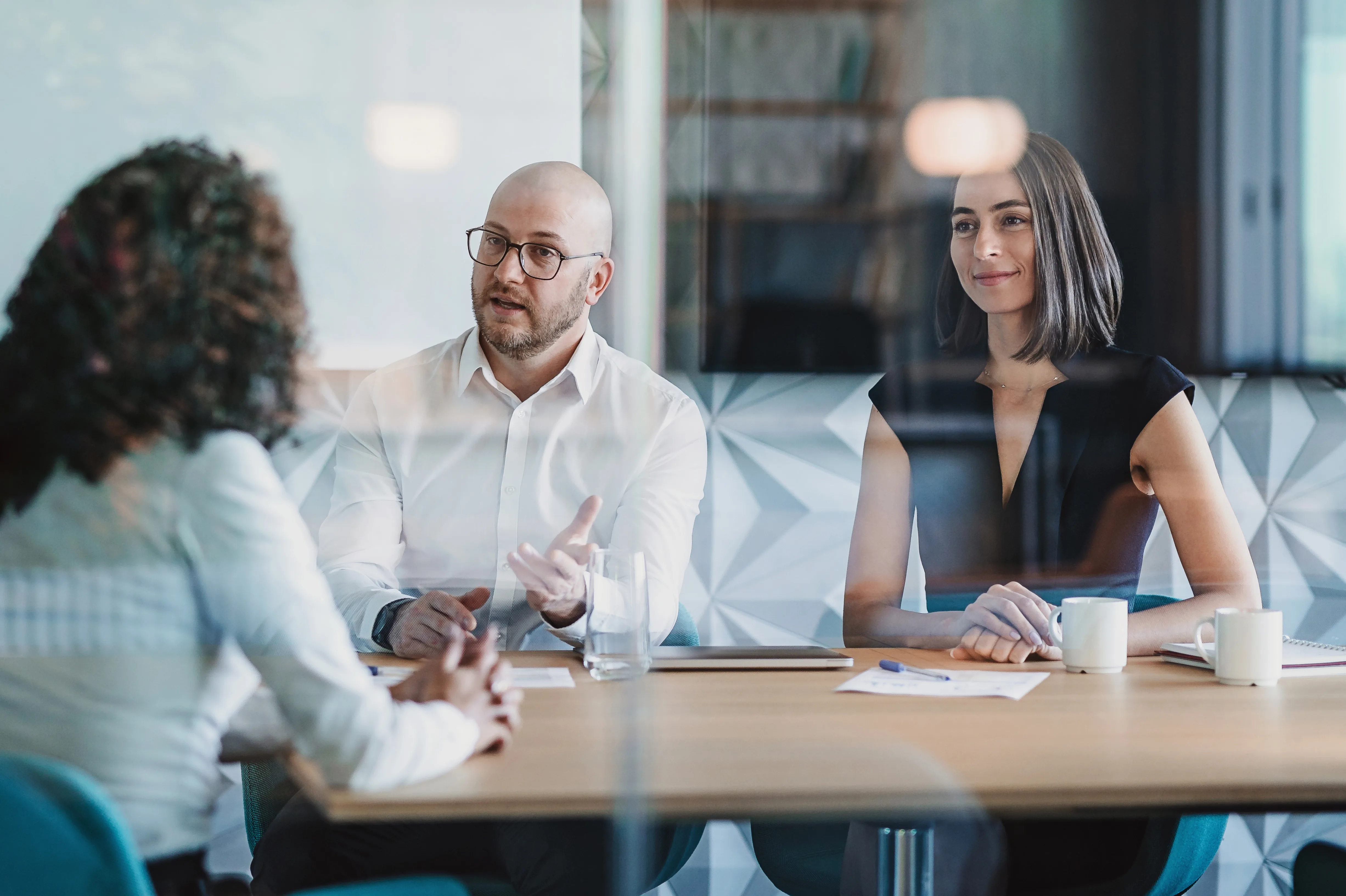 Three colleagues talk across a conference table in a bright glass-walled office