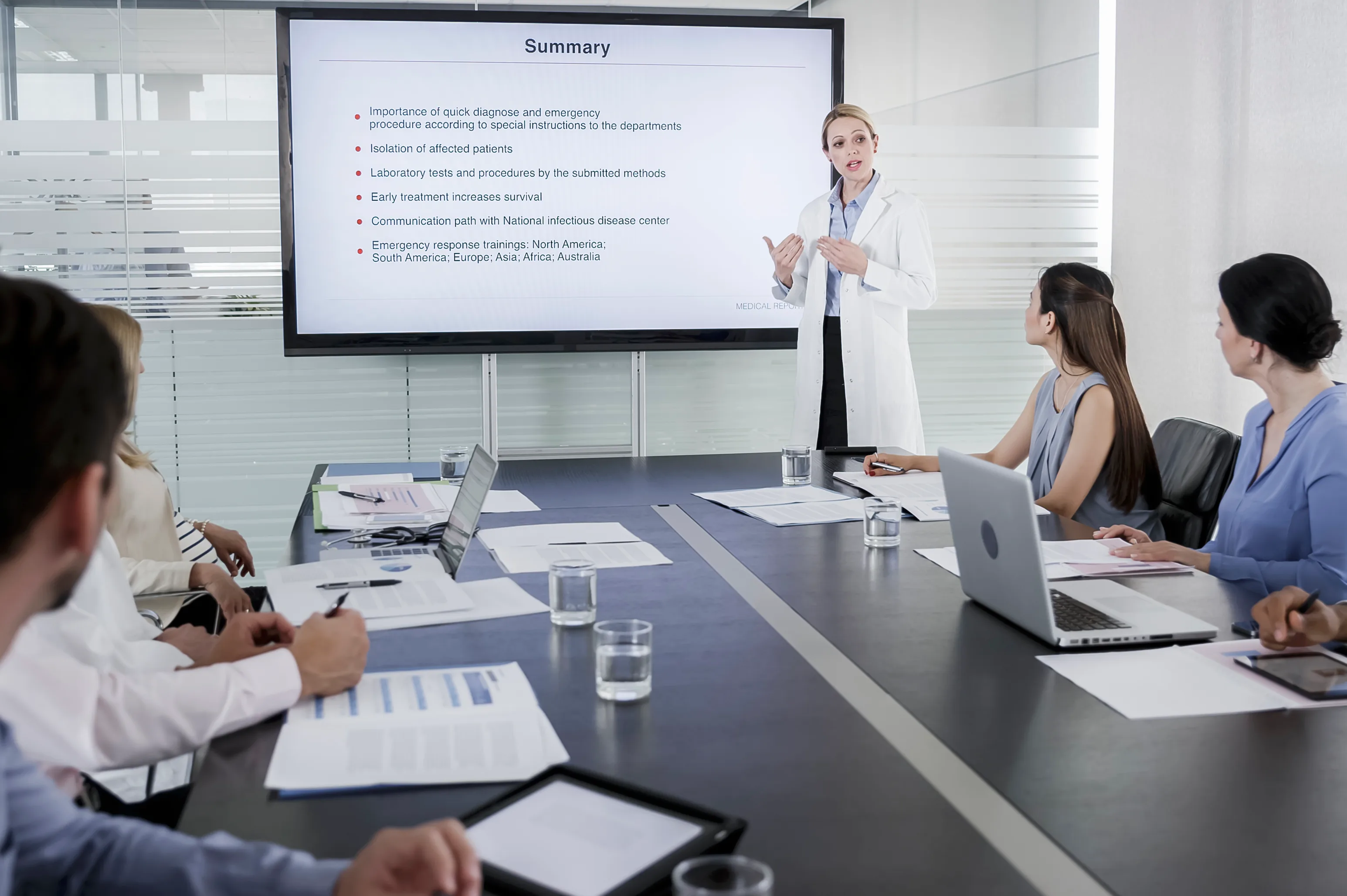 Woman in a lab coat presents a summary slide to seated colleagues in a boardroom