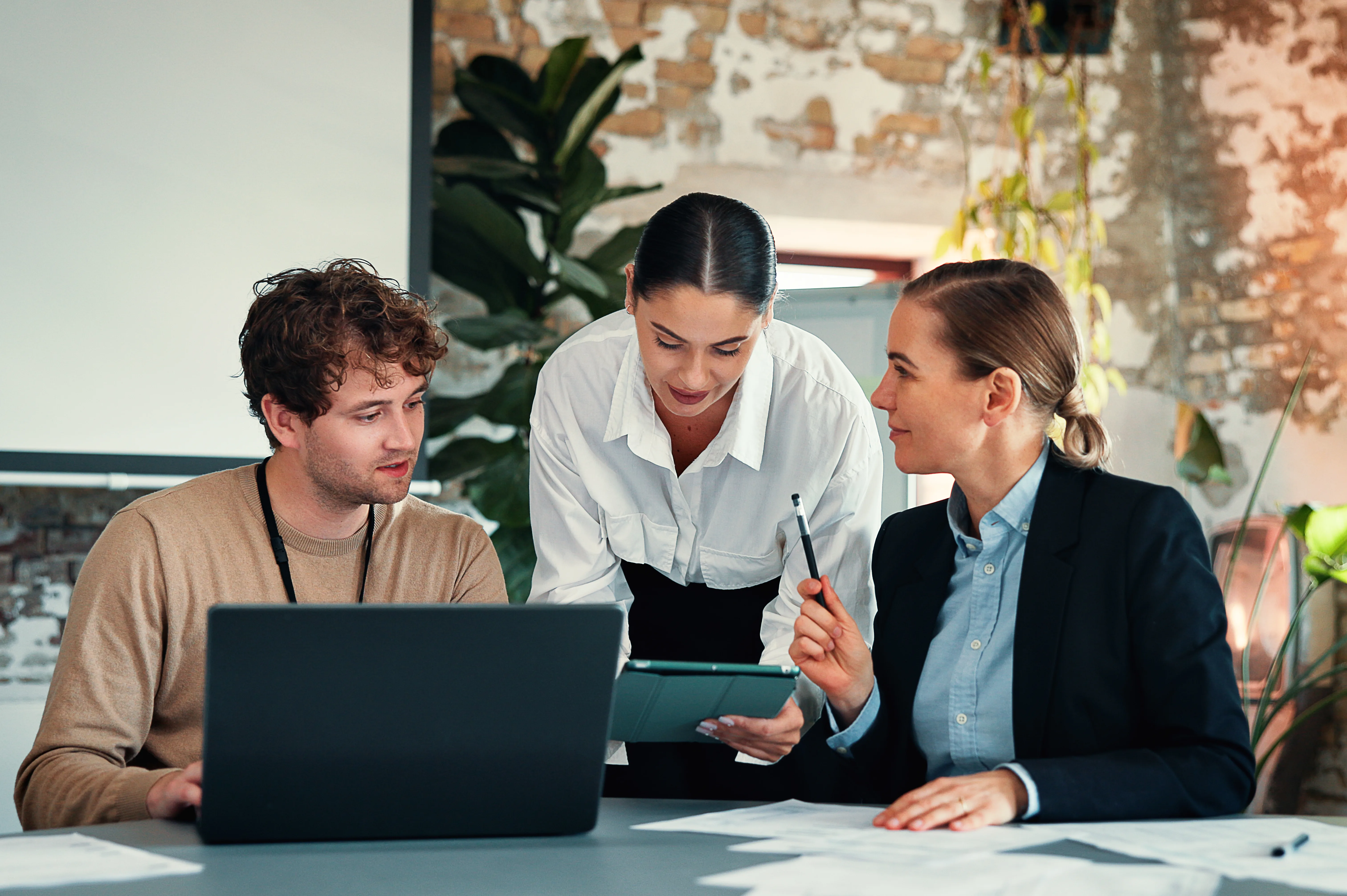 Three colleagues review a laptop and tablet together at a desk in a loft office