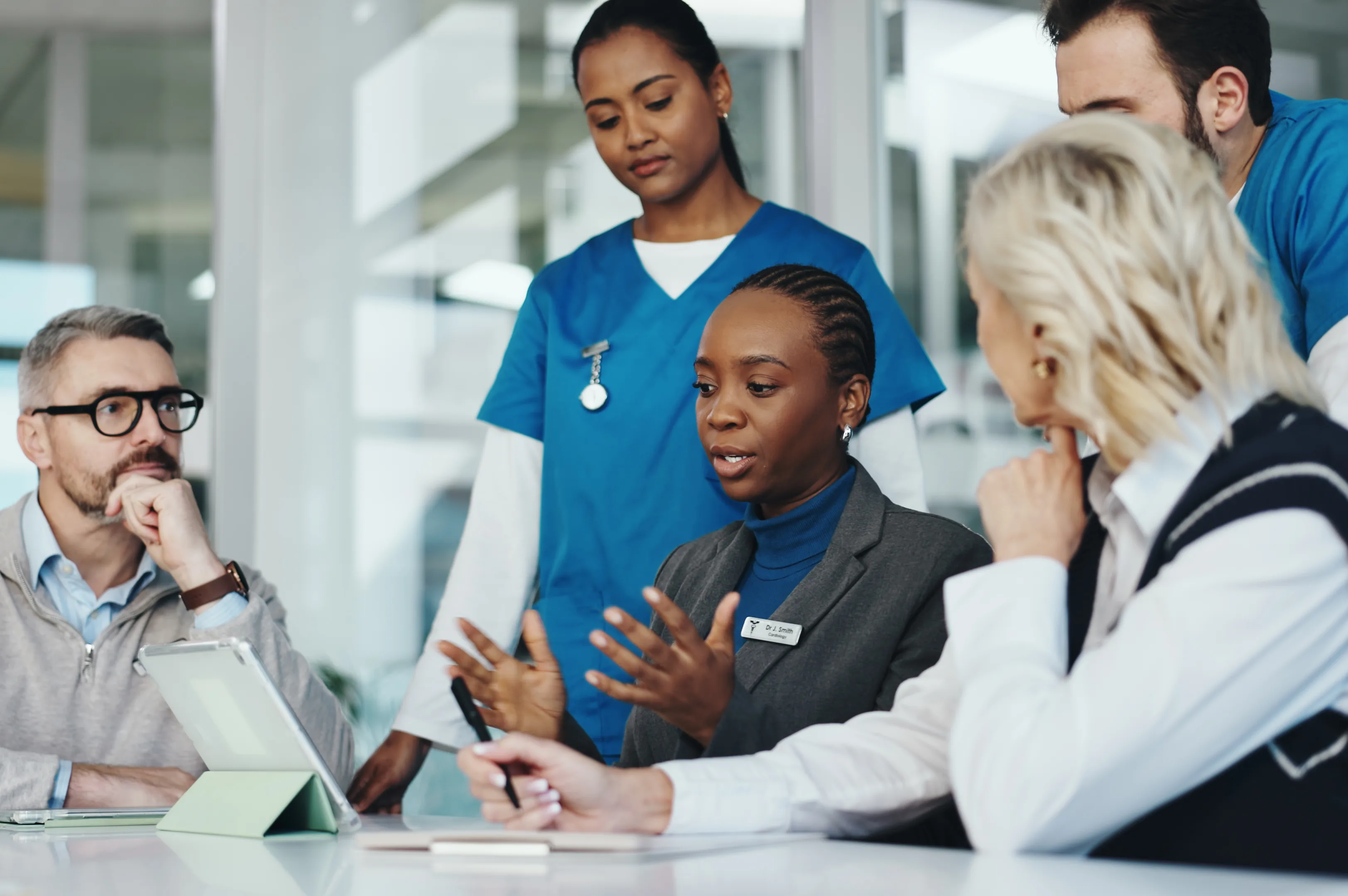 Clinicians in scrubs and a suit discuss around a table with a tablet in a hospital