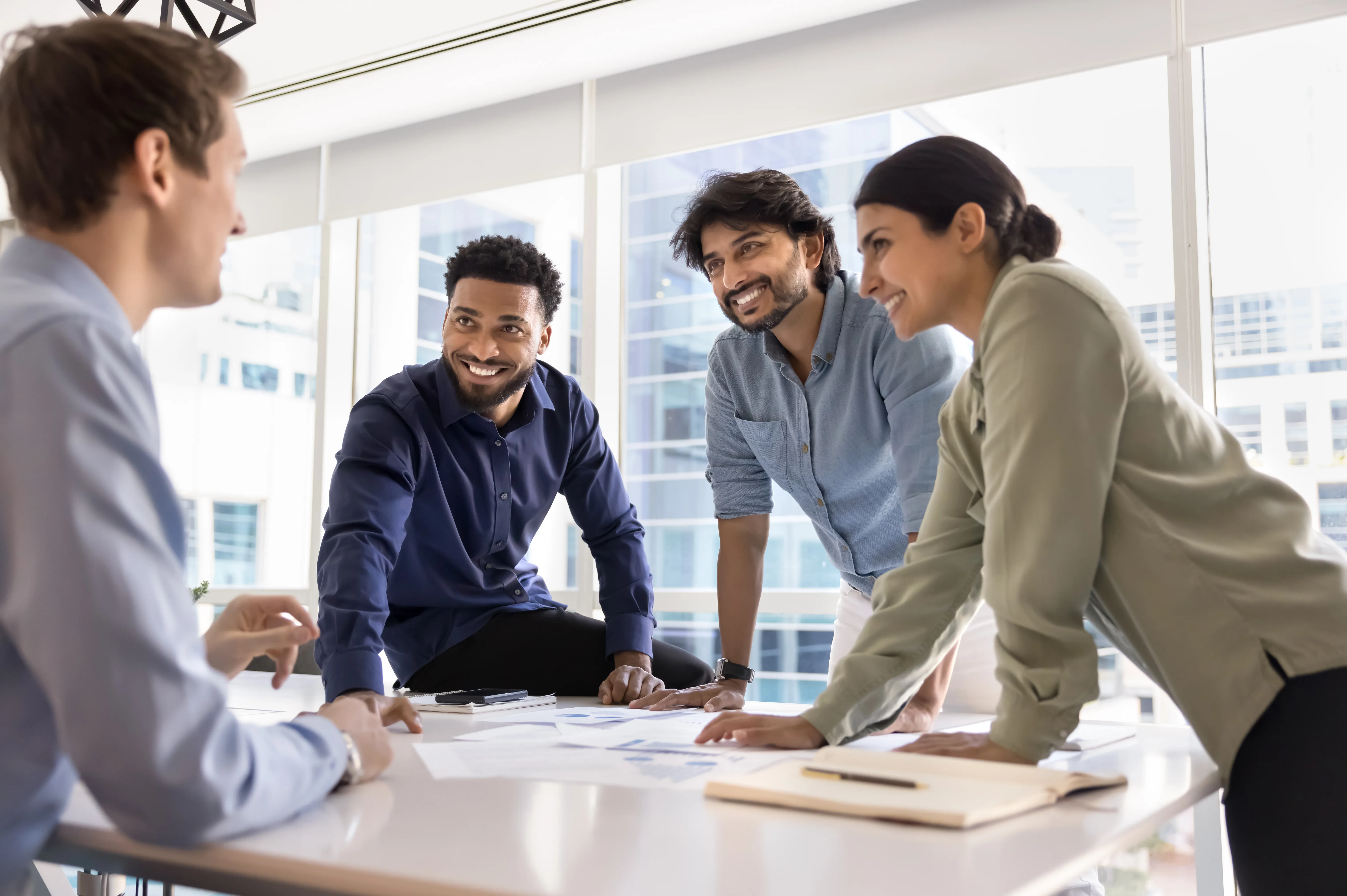 Four coworkers smile and talk while leaning on a table by office windows