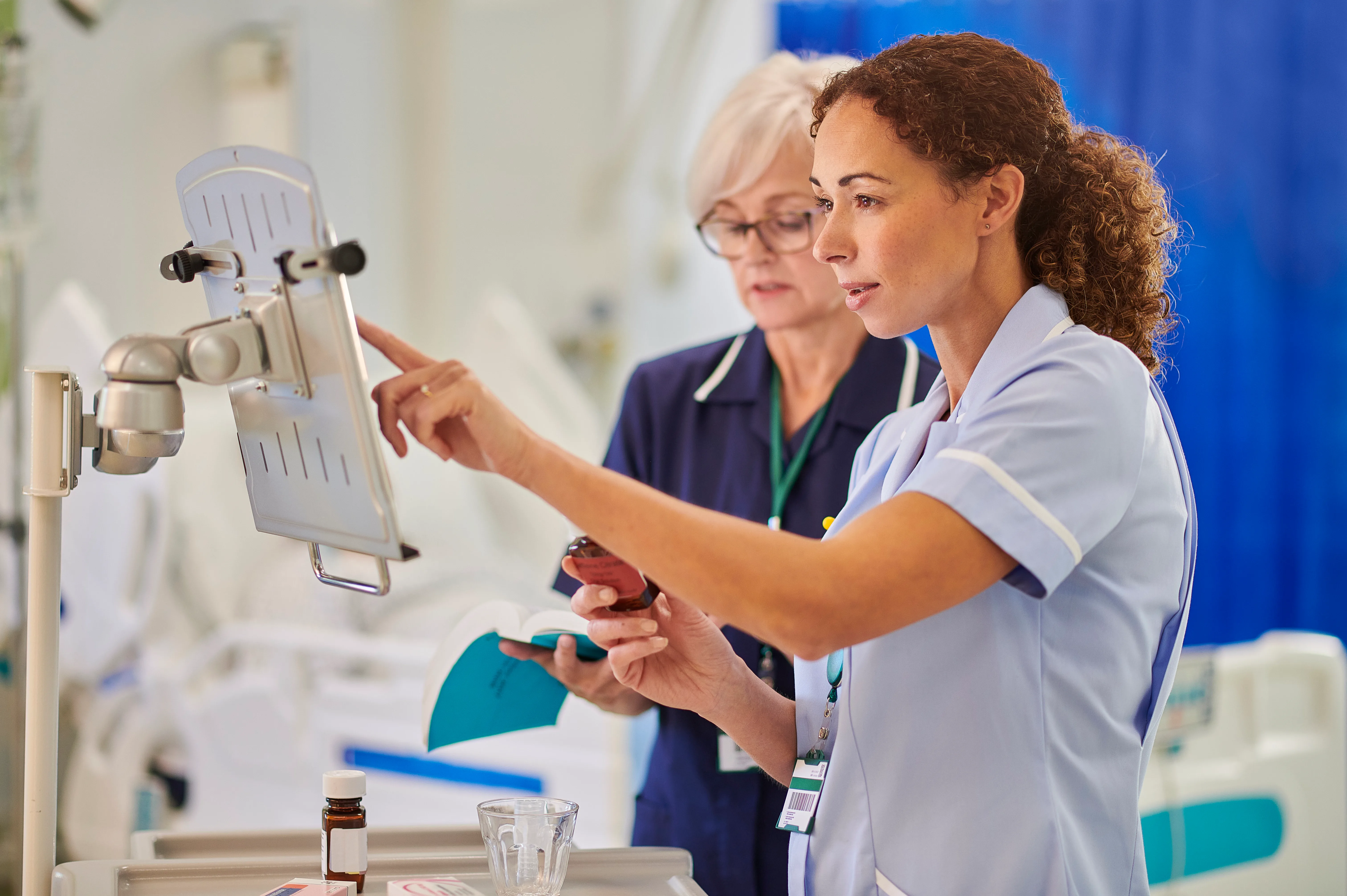 Two nurses in uniform review a medication bottle beside a monitor in a hospital ward