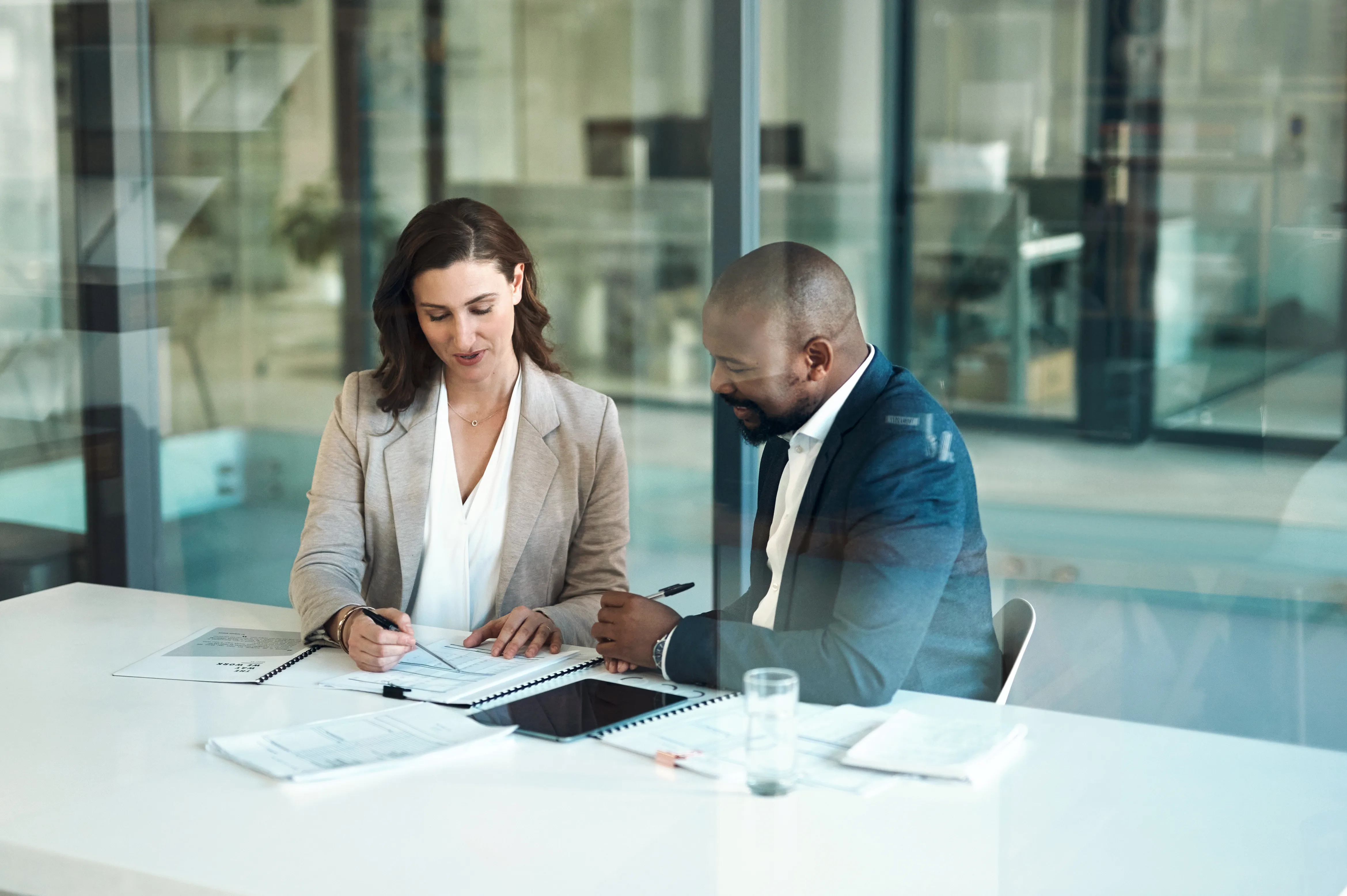 A woman and a man review documents together at a white table behind glass