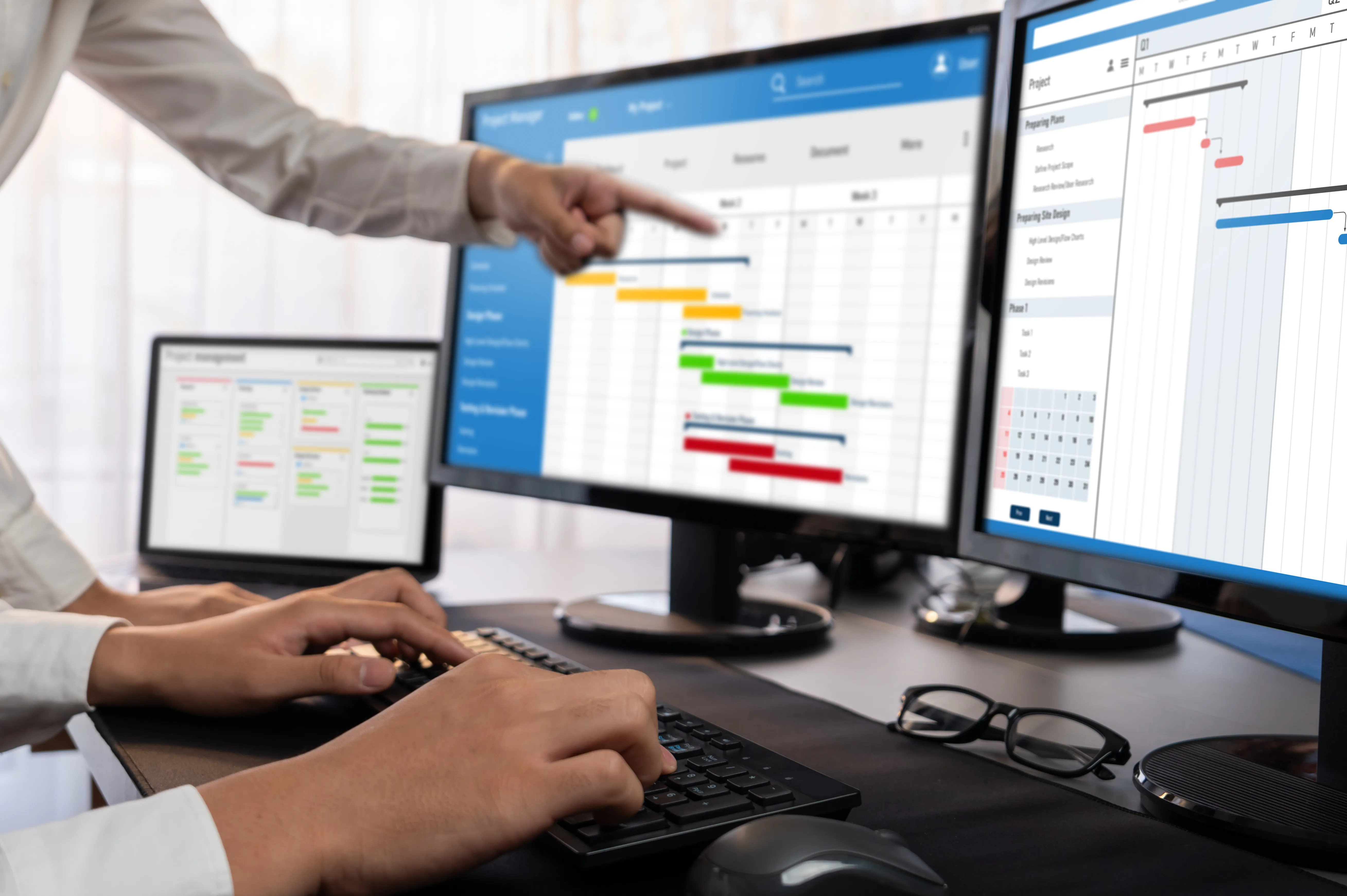 Person points at project timeline Gantt charts on dual monitors while another types