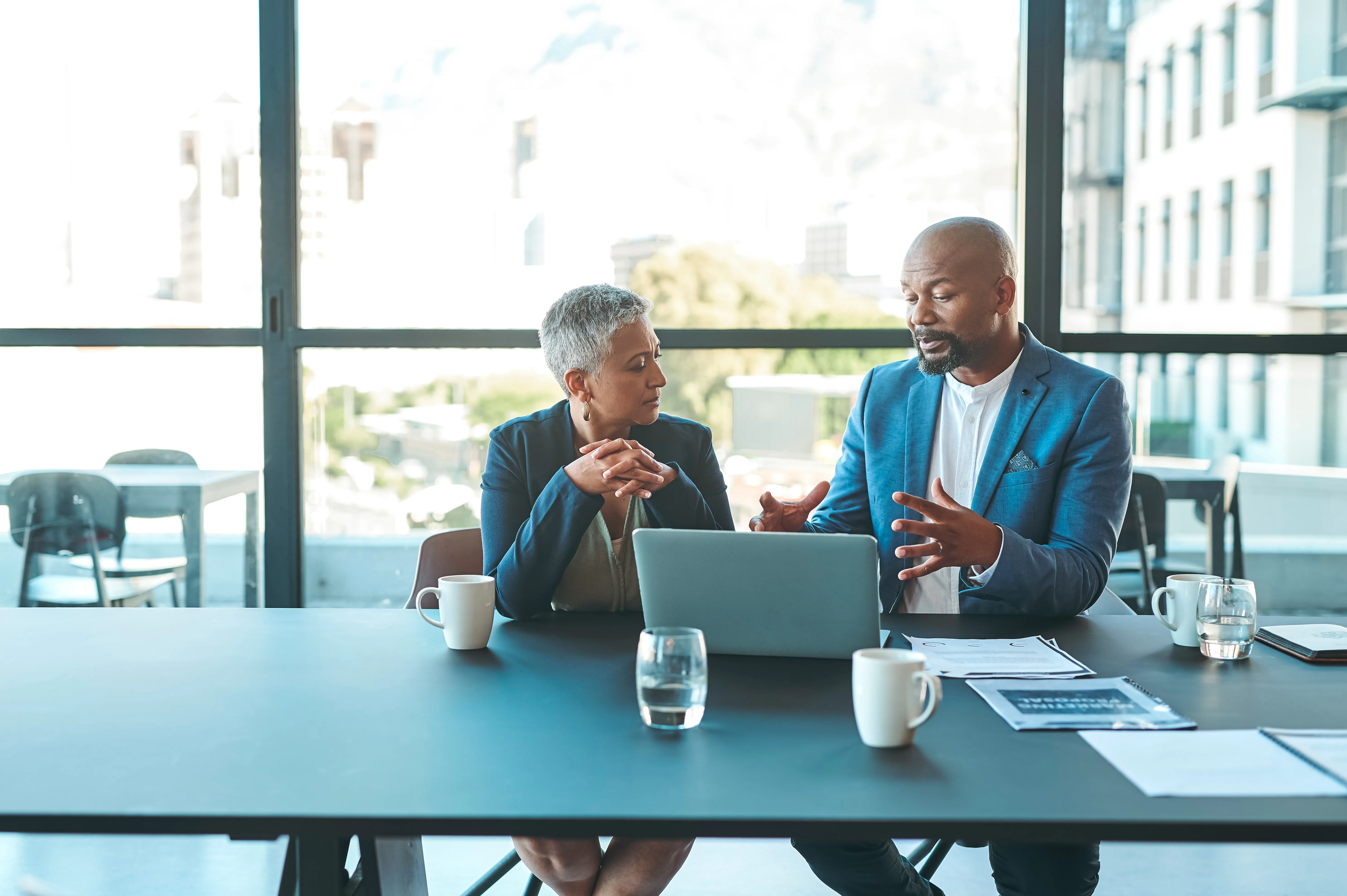 A woman and a man in a blazer discuss over a laptop by large office windows