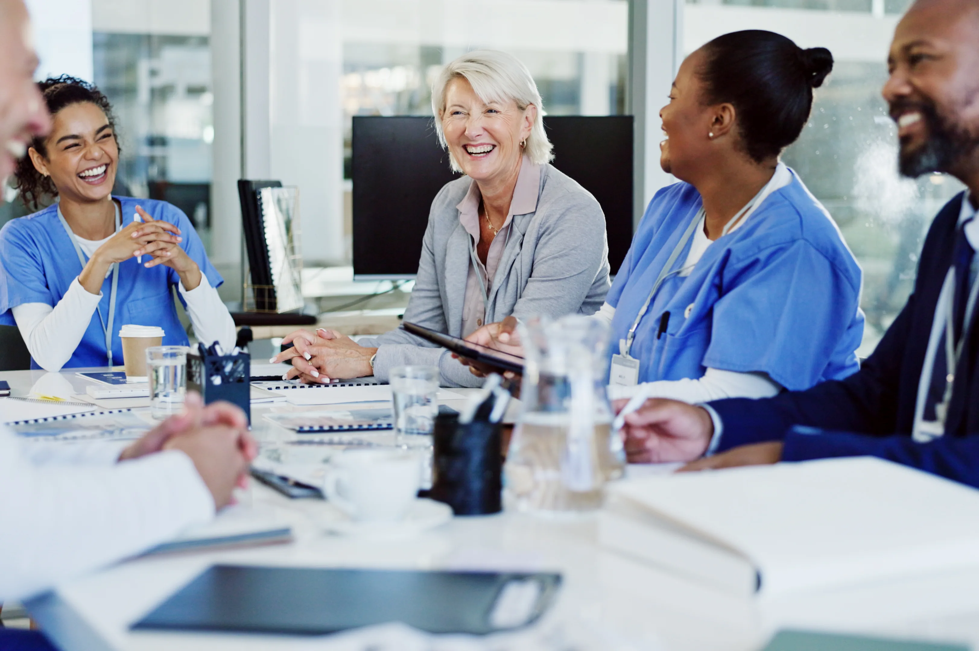 Nurses in scrubs and colleagues laugh together around a boardroom table