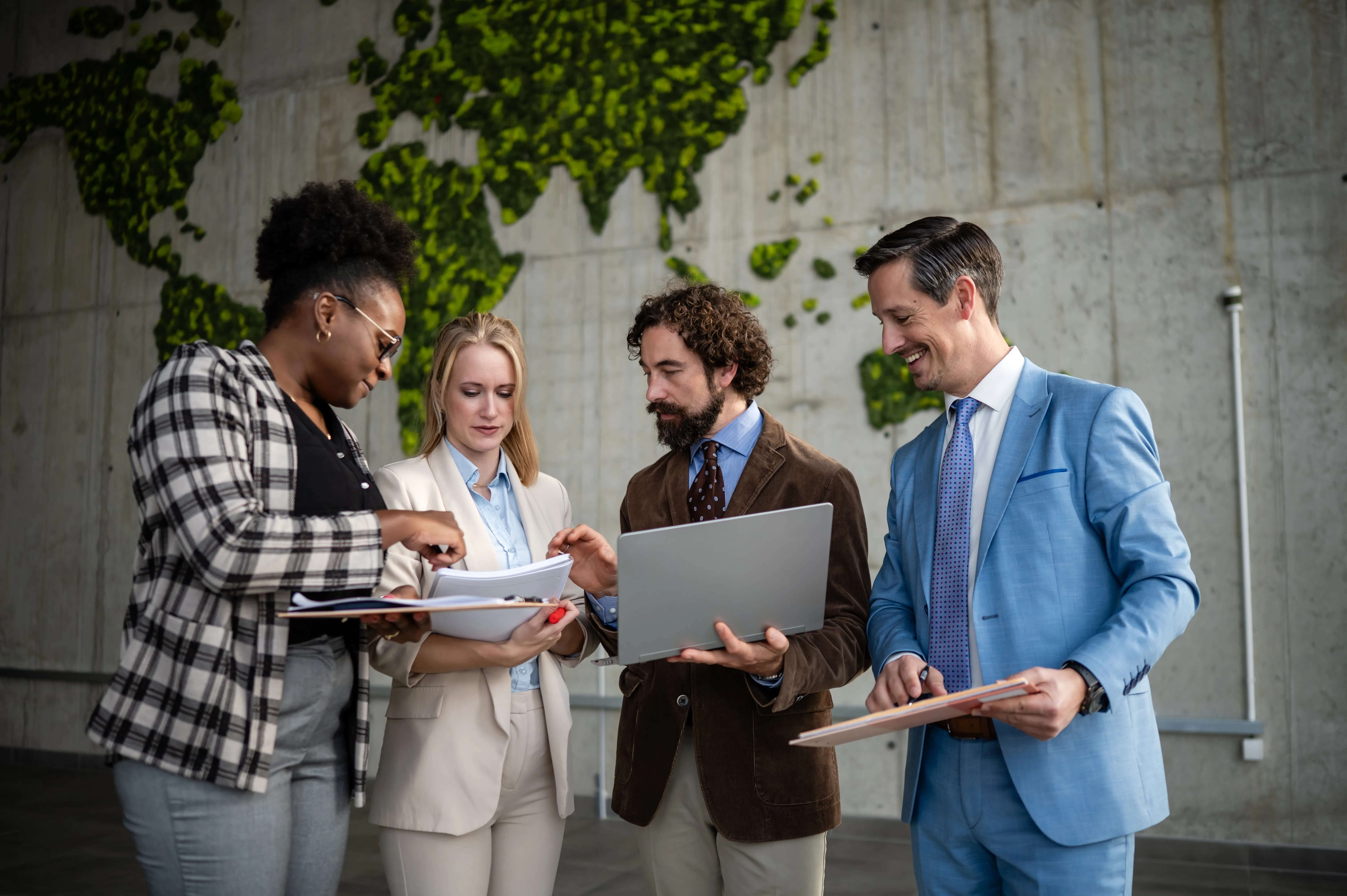Four professionals review a laptop and papers before a green moss world map wall