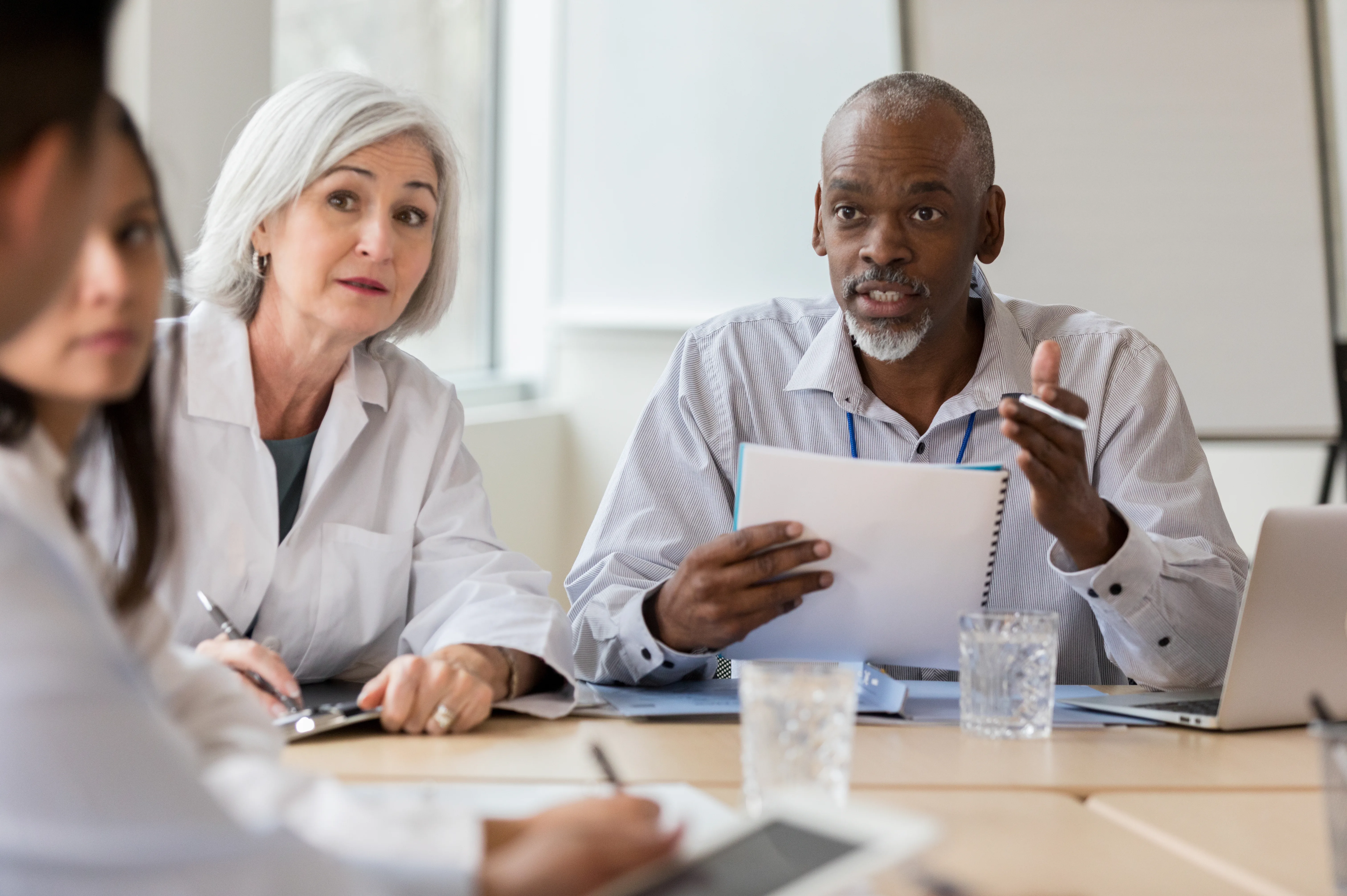 An older man gestures with papers while a woman listens across a meeting table