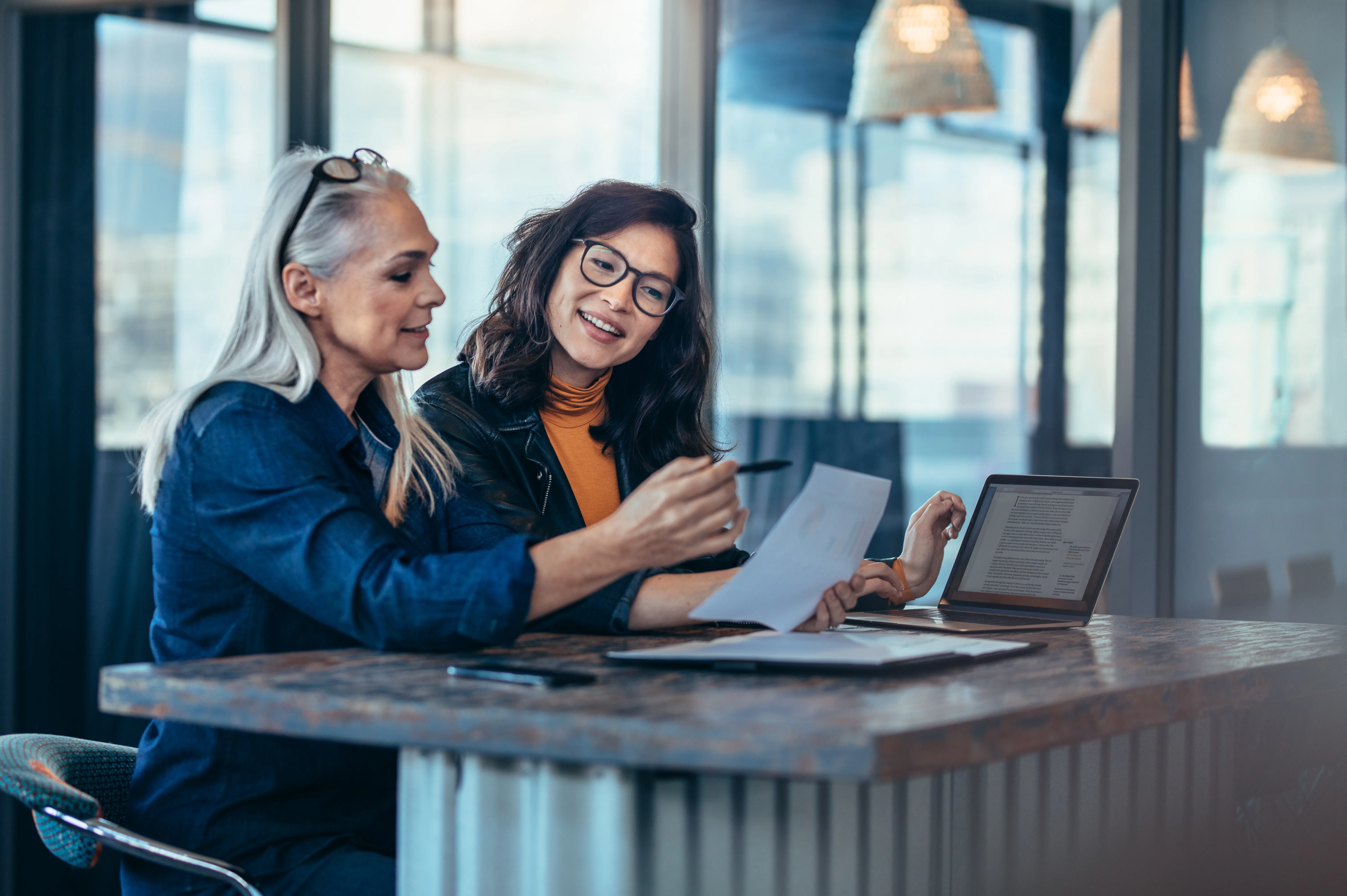 Two women review a document and laptop together at a high table by windows